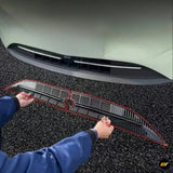 Black and red vehicle grille insert being positioned on a car bumper by a person wearing blue gloves and a jacket