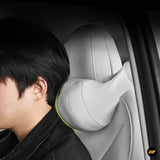 Grey car seat neck support cushion fitted around a headrest, shown beside a seated driver in a vehicle interior