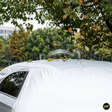 Silver car cover protecting a vehicle outdoors beneath leafy trees, with fallen leaves resting on the covered roof