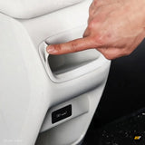 Hand pointing to a recessed storage compartment in a white vehicle interior beside a small black control panel