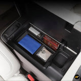 Open centre console storage box in electric vehicle showing organised compartments with wallet, tissue, and cigarette pack inside