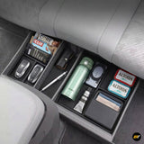 Underseat car organiser tray with compartments holding snacks, keys, water bottle, charger, and wallet inside a grey vehicle interior.