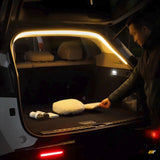 Person placing items in a car boot illuminated by warm white LED light strips around the trunk opening at night